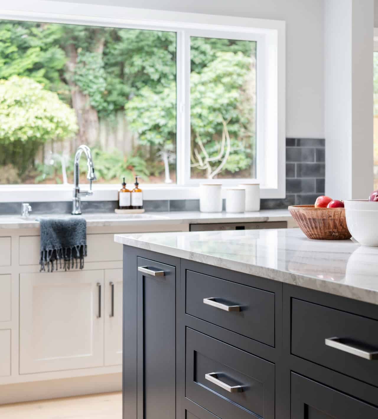 Modern kitchen with white cabinets, gray island, and large window showing greenery.