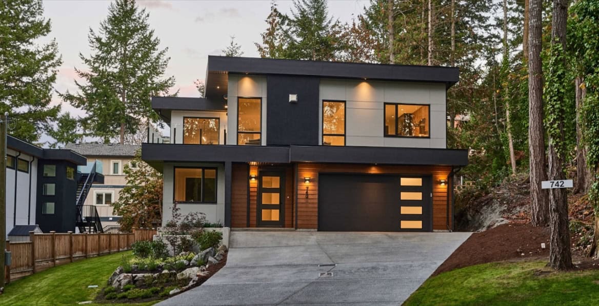 Modern two-story home with dark exterior, large windows and a double garage in a wooded setting.