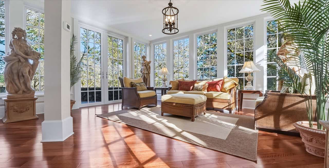 Bright sunlit living room with large windows, wooden floors, and elegant seating in a spacious home.