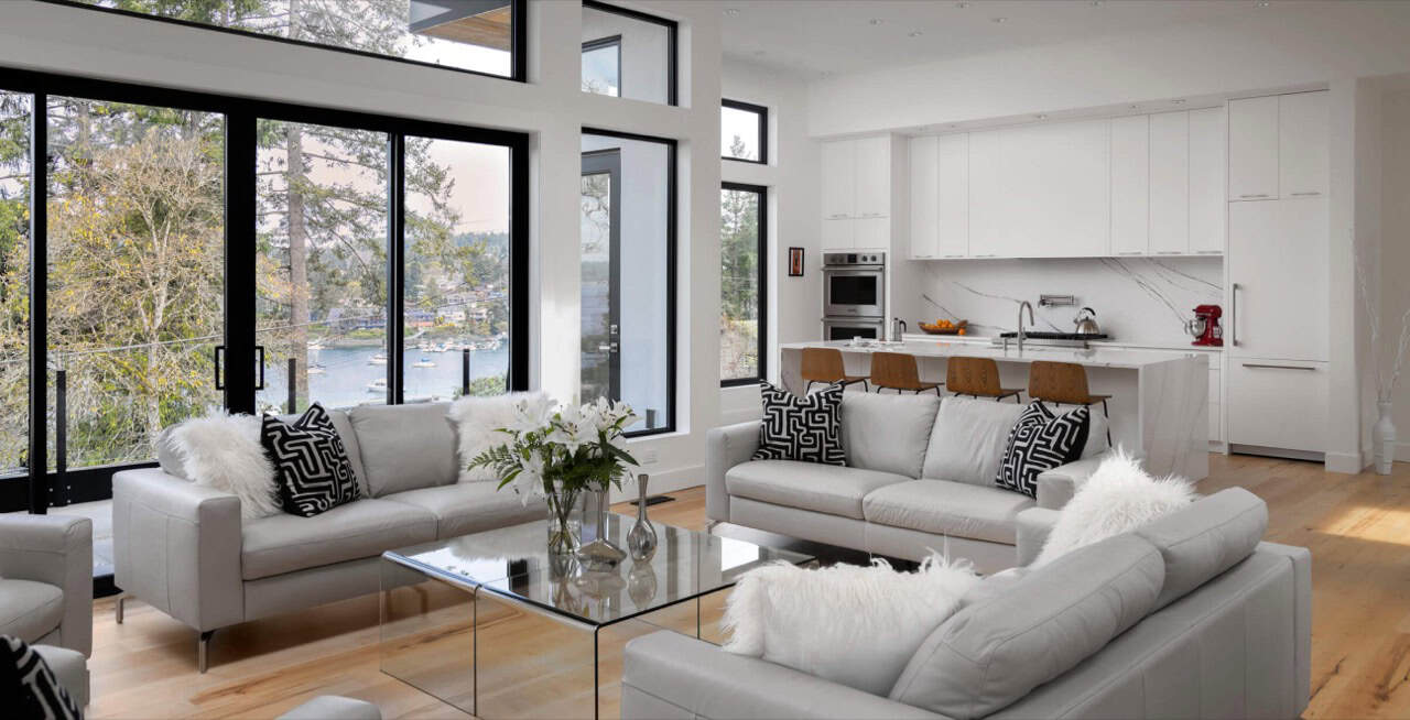 Modern living room with large windows, white furnishings, and a kitchen in the background.