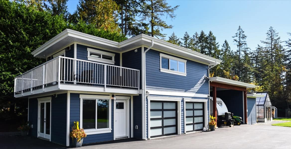 Blue modern two-story home with white trim, large balcony and attached garage in a wooded setting.