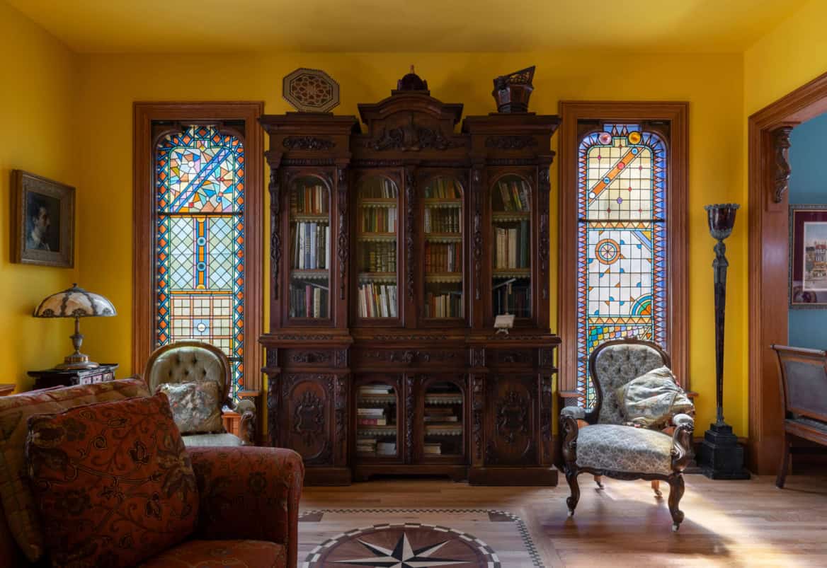 Victorian library with ornate wooden cabinet, stained glass windows, and vintage chairs.