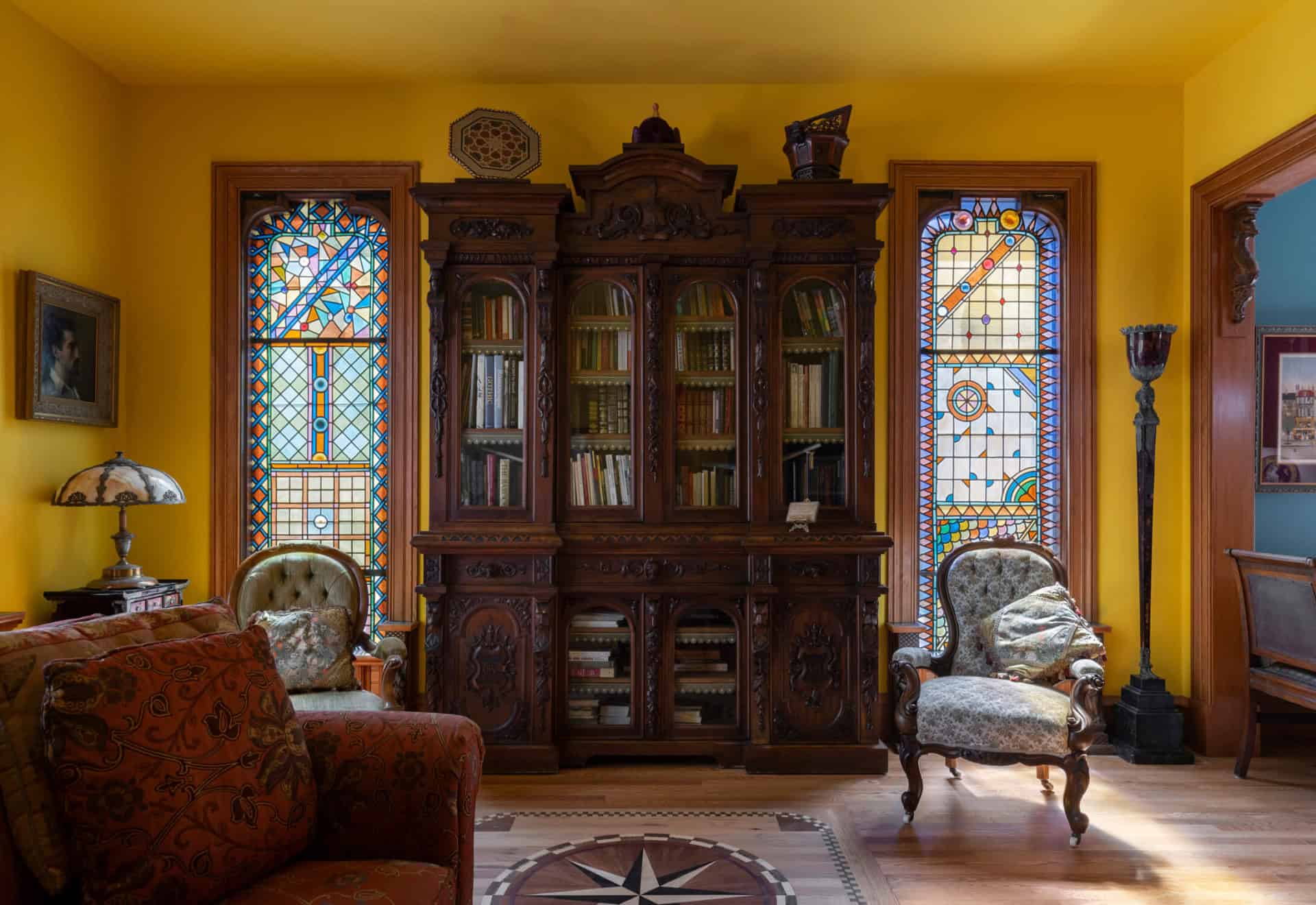 Victorian library with ornate wooden cabinet, stained glass windows, and vintage chairs.