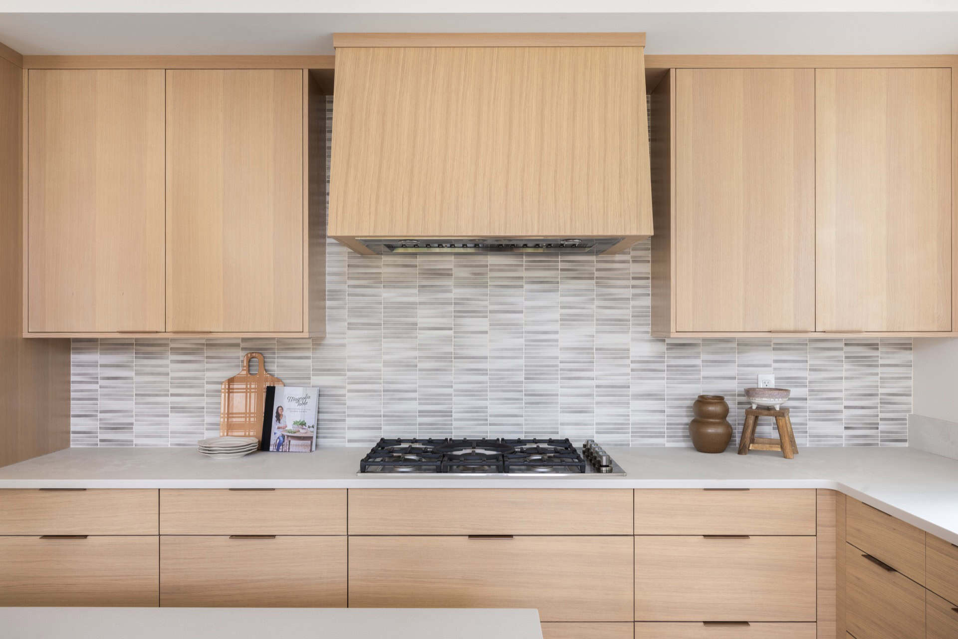 Bright modern kitchen with light wood cabinets and gas stove under a range hood.
