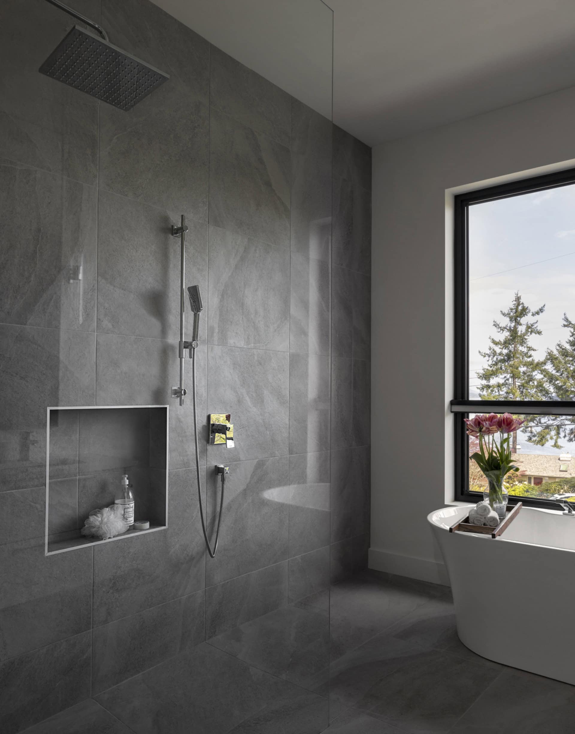 Modern gray-tiled bathroom with glass shower, freestanding tub, and large window overlooking trees.