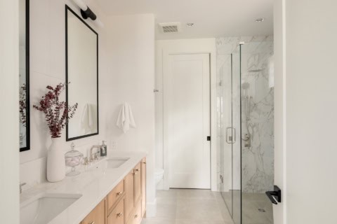 White contemporary bathroom with double vanity, marble wall accents, and glass shower enclosure.
