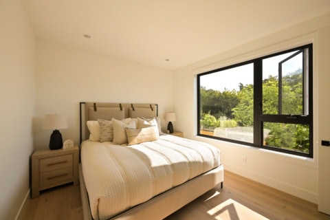 Bright airy bedroom with large window overlooking greenery and a neutral-toned bed.