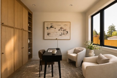Interior view of a modern home office with beige seating, wooden cabinets, and large window showing庭.