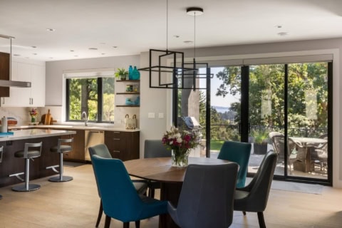 Interior of a modern kitchen and dining area with large glass doors to a deck and greenery outside.