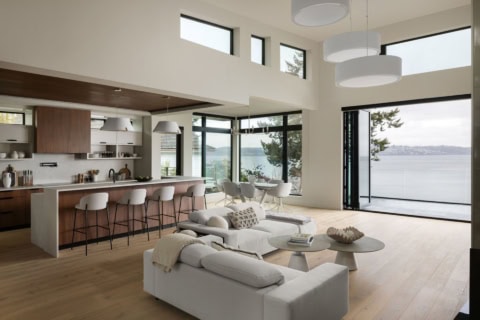Bright open-concept living area with large windows and waterfront view, modern kitchen and white/cre.