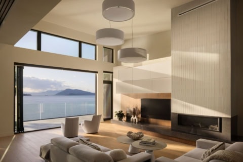 Open living room with large windows overlooking water and distant island, modern fireplace, and airy.