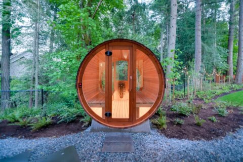 Round wooden barrel sauna surrounded by green trees in natural garden.