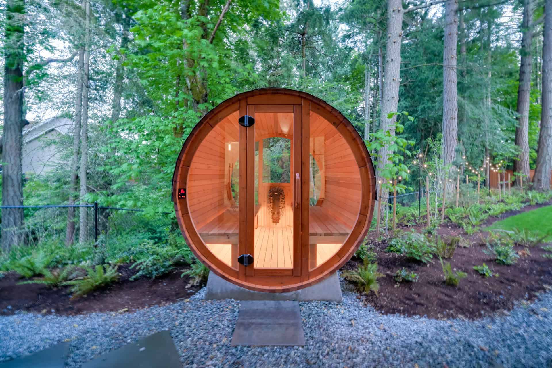 Round wooden barrel sauna surrounded by green trees in natural garden.
