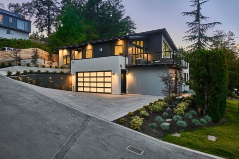 Modern Victoria BC custom home with expansive driveway and illuminated garage doors at dusk.