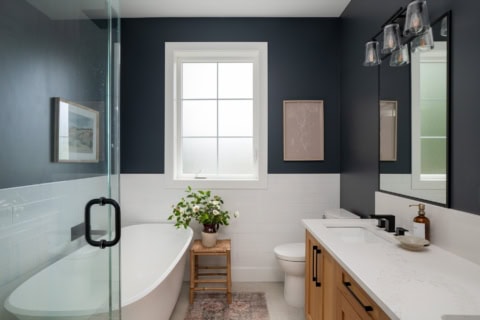 Modern bathroom with dark blue walls, white tile, freestanding tub, and wooden vanity.