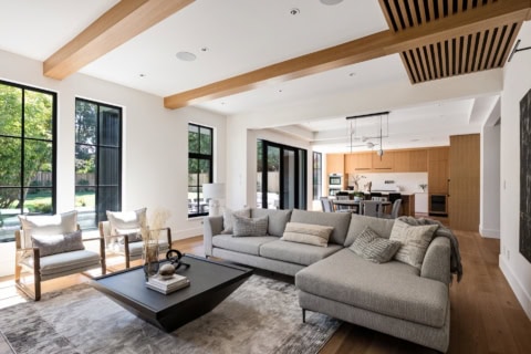 Open-plan living room with large windows, gray sofa, wooden beams, and contemporary kitchen in the背景.