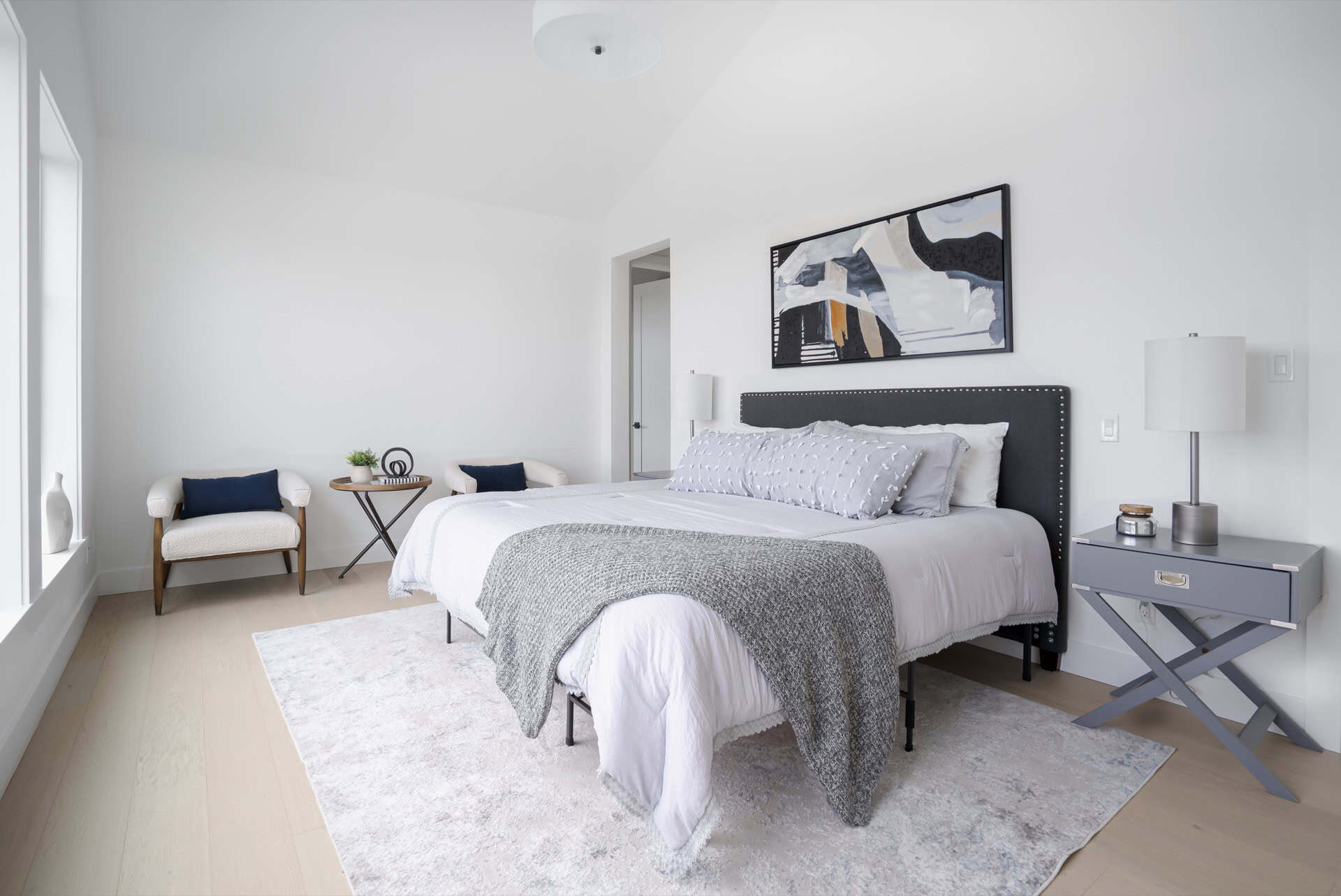 Bright modern bedroom with a gray upholstered bed, matching nightstands, abstract wall art, and a he.