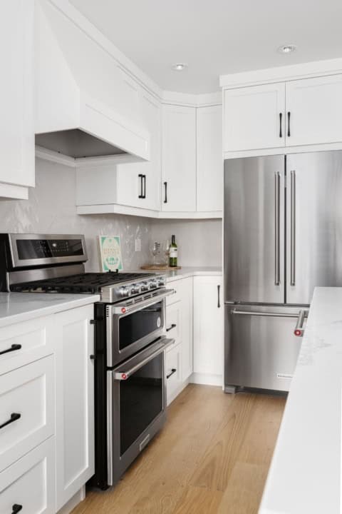 Modern white kitchen with stainless steel fridge and range, light wood floor.