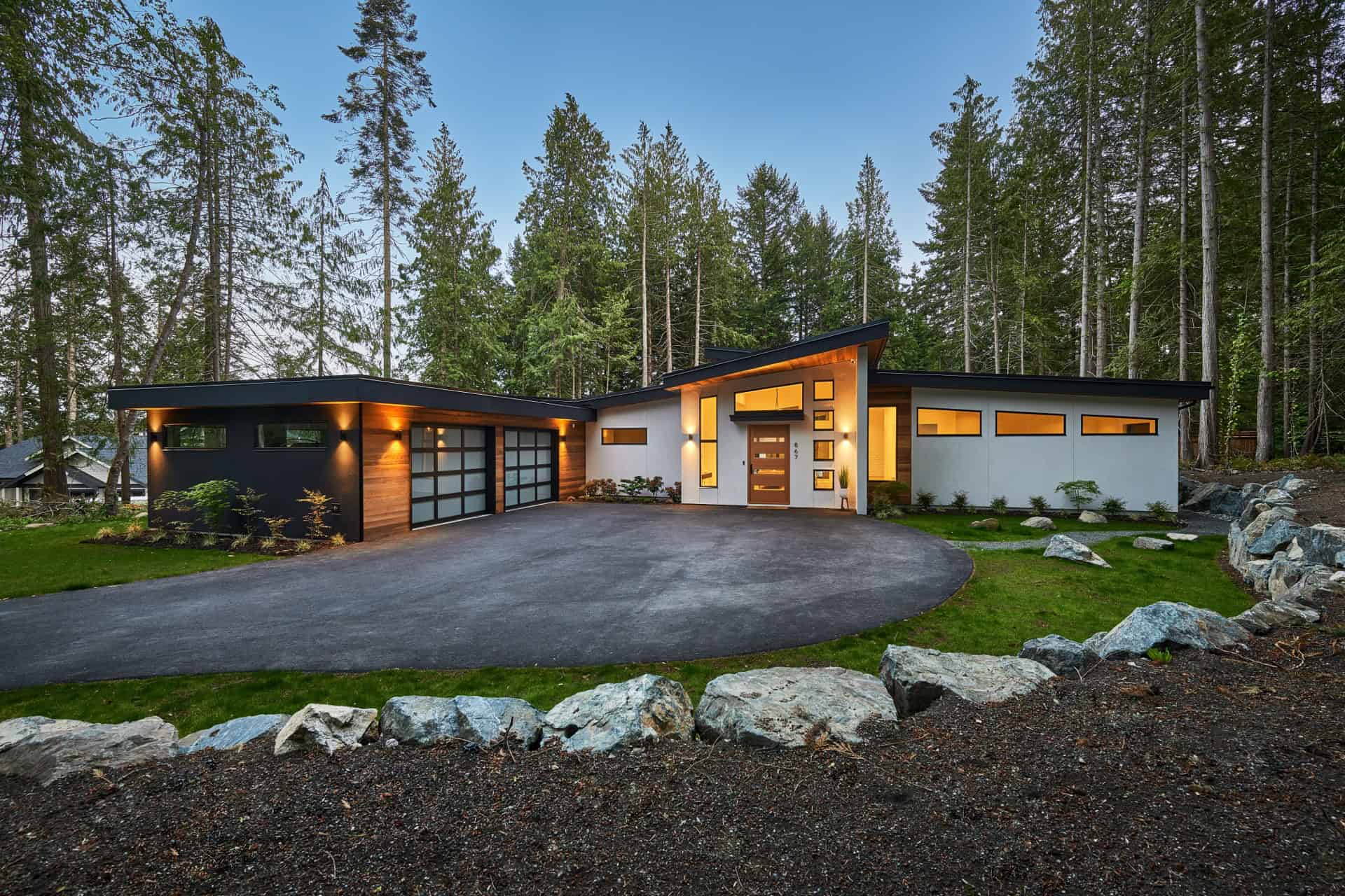 Modern single-story home by a forested setting with a curved driveway and stone landscaping.