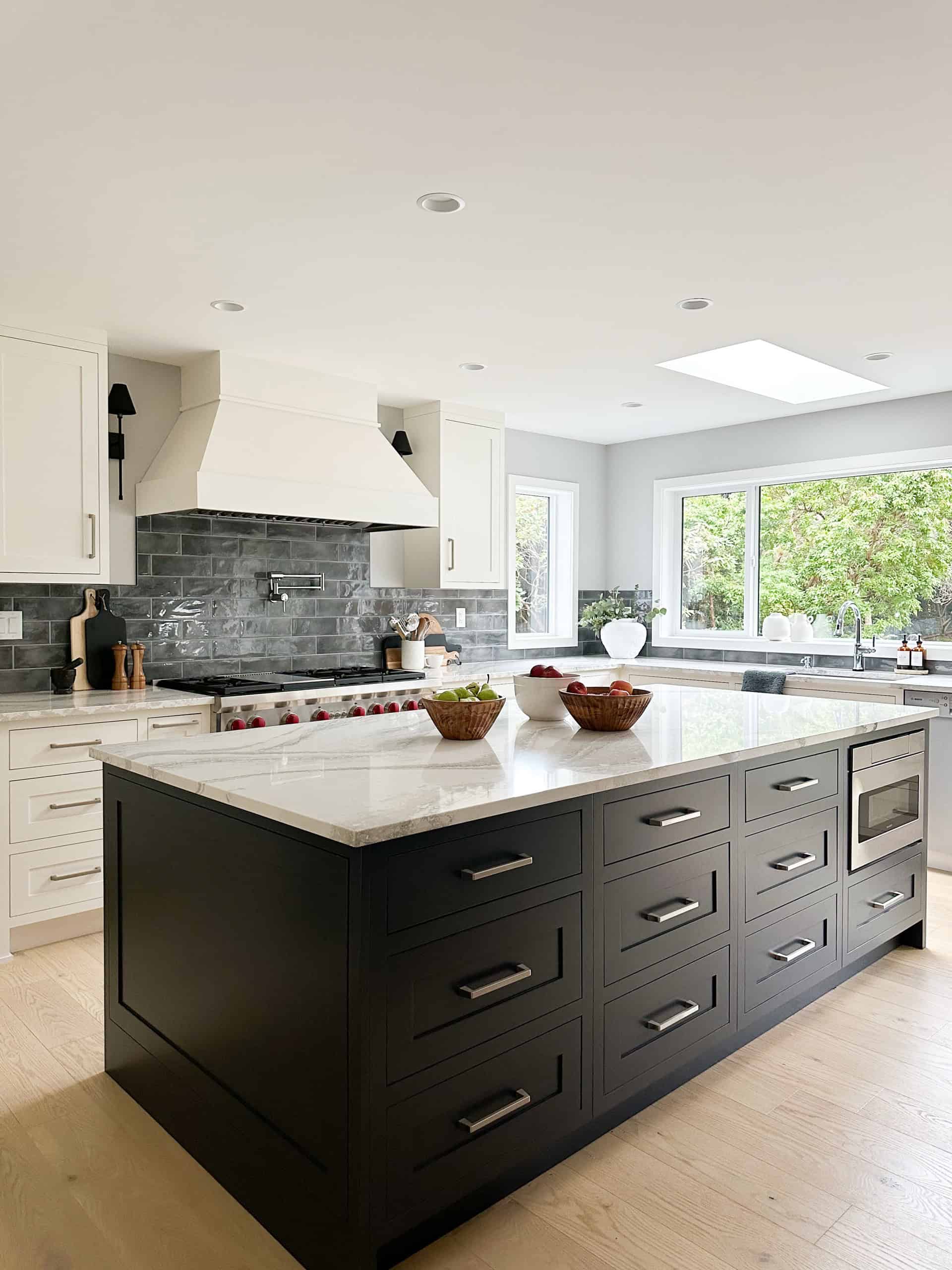 Modern kitchen with an oversized dark island and white cabinetry, bright natural light, and a brick-.