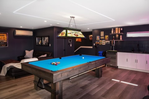 Modern game room with a blue pool table and neon lighting, sleek dark walls, and a kitchenette in a.