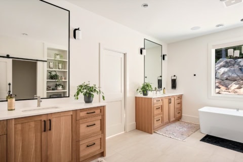 Bright modern bathroom with dual wooden vanities, wide mirrors, potted plants, and a freestanding so.